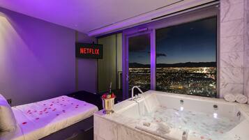 A hotel room with a bed, a large window with a city view, a bathtub, and a sign that says 'NETFLIX'.
