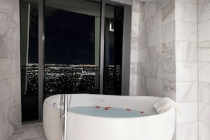 A modern bathroom with a large bathtub, marble walls, and a city view through large windows.