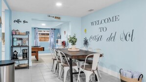 Un coin repas avec une table en bois et des chaises blanches, un bar à café équipé de divers appareils pour préparer le café, et un mur orné d’une enseigne indiquant « Bienvenue à la casa del sol ».