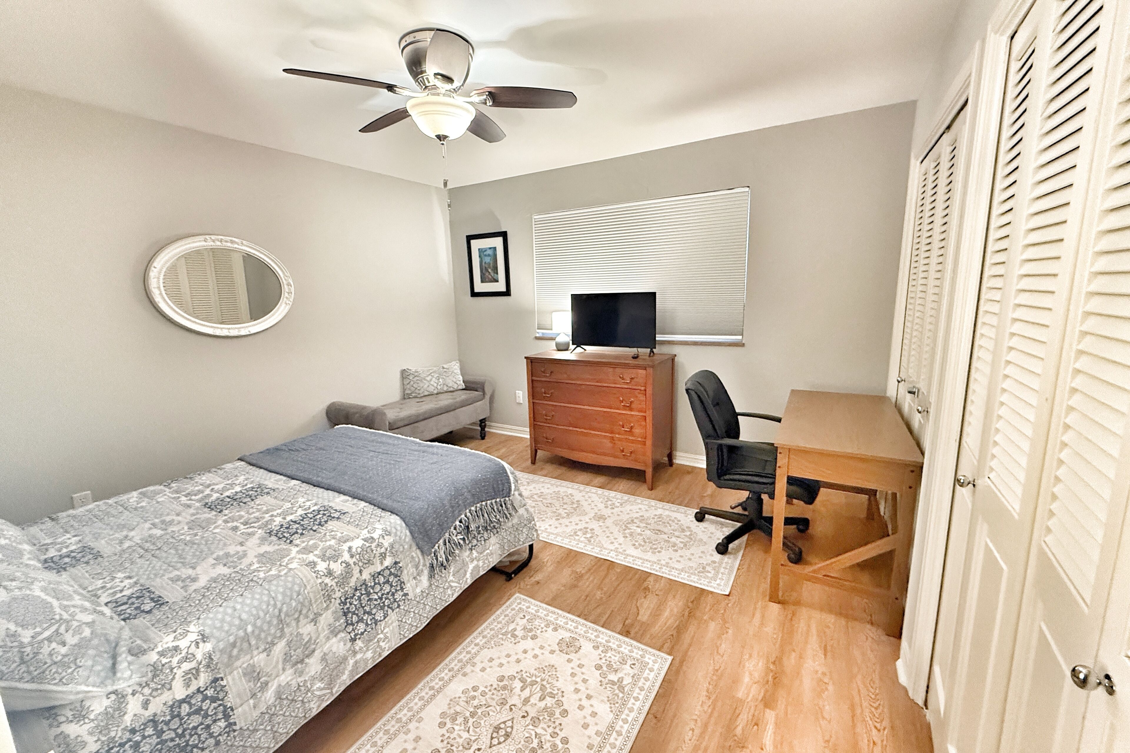 Une chambre à coucher comprenant un lit, un bureau, une chaise, une commode, un ventilateur de plafond, une fenêtre avec des stores et une porte dotée de volets à persiennes.