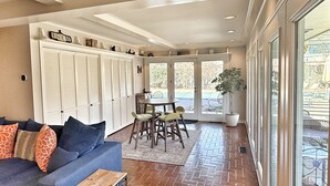 A spacious living area with a blue sofa, a wooden coffee table, and a dining table with chairs. There is a large window with a view of the outdoors, and a potted plant near the window.