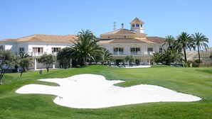 A golf course with a large sand bunker, palm trees, and a building with a tiled roof and balconies. - Manilva Gardens G.A6 (Manilva)