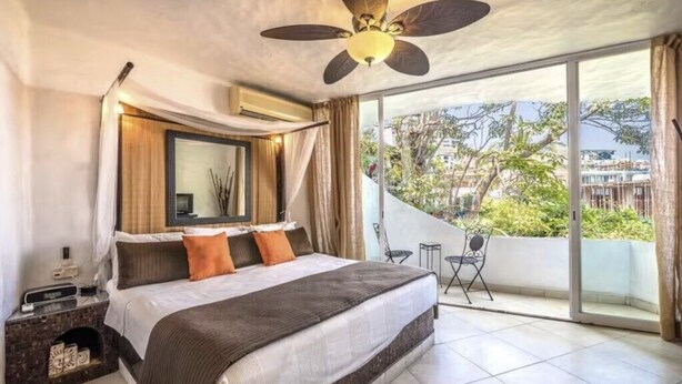 Un dormitorio con una cama grande, ventilador de techo, balcón con vistas y una pequeña mesa con sillas.
