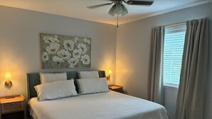 Et soveværelse med en seng, to sengeborde, en ventilator i loftet, en vægmonteret lampe, et vindue med gardiner og et blomsterbillede.