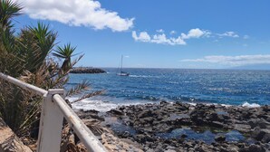 Una costa rocosa con cielo azul claro y un velero a lo lejos.