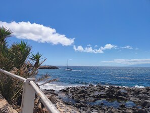 Una costa rocosa con cielo azul claro y un velero a lo lejos.