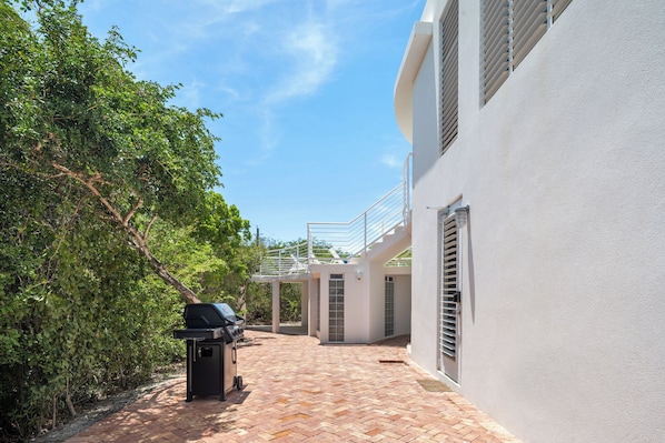 En overdækket terrasse med grill, en hvid bygning med balkon, og en brostensbelagt sti.