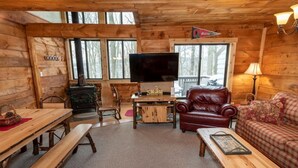 Ein gemütliches Blockhaus-Interieur mit Holzvertäfelung, einem Fernseher, einem Ledersessel, einer Couch und einem Esstisch mit Bänken.