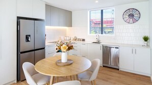 A modern kitchen with a stainless steel refrigerator, a round wooden table with white chairs, a vase with flowers, and a clock on the wall.