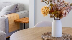 A modern living room with a round wooden table, a white vase with flowers, a grey sofa, and a large abstract painting on the wall.