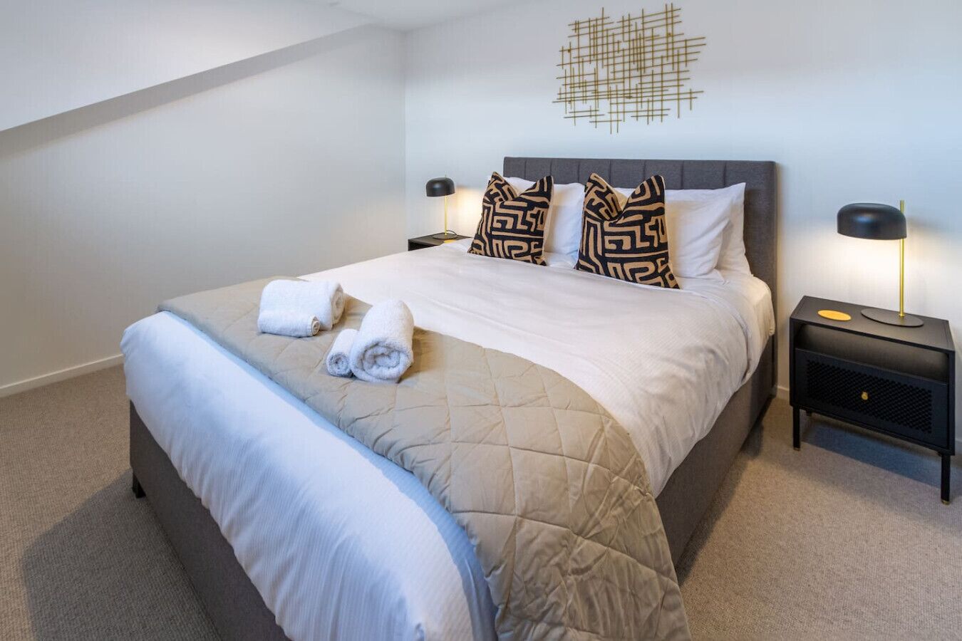 A neatly made bed with a quilted duvet, two pillows with a geometric pattern, and two bedside lamps on a nightstand.