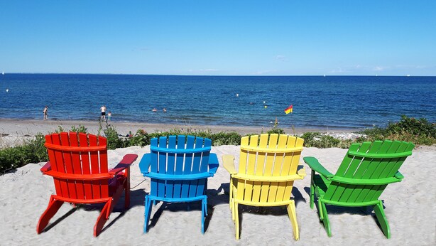 Quatre chaises Adirondack de couleurs rouge, bleue, jaune et verte, placées sur une plage de sable, avec l’océan en arrière-plan.