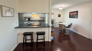 A modern kitchen with a central island, two bar stools, a dining area with a table and chairs, and a hallway leading to another room. - Peaceful Hyde Park Condo Near UT/Moody with Free Parking, Fast WiFi, King Bed (Austin)