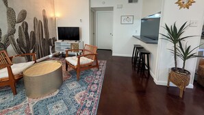 A living room with a flat-screen TV, two wooden chairs, a patterned rug, and a cactus wall art piece. - Peaceful Hyde Park Condo Near UT/Moody with Free Parking, Fast WiFi, King Bed (Austin)