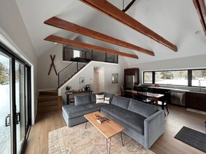 Una sala de estar moderna con un sofá modular, vigas de madera, una escalera y una zona de cocina.