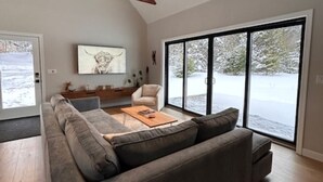 Un salon moderne avec un canapé d’angle, une table basse et une grande fenêtre donnant sur des arbres couverts de neige.