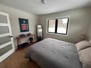 A bedroom with a bed, a window, a mirror, a shelf with items, and a door.