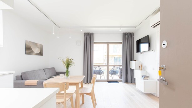 A modern, well-lit room with a dining area, a sofa, and a TV mounted on the wall. - New 2025 Pervalka Neringa Flat White Dunes: AC, Heating, Private Balcony (Pervalka)