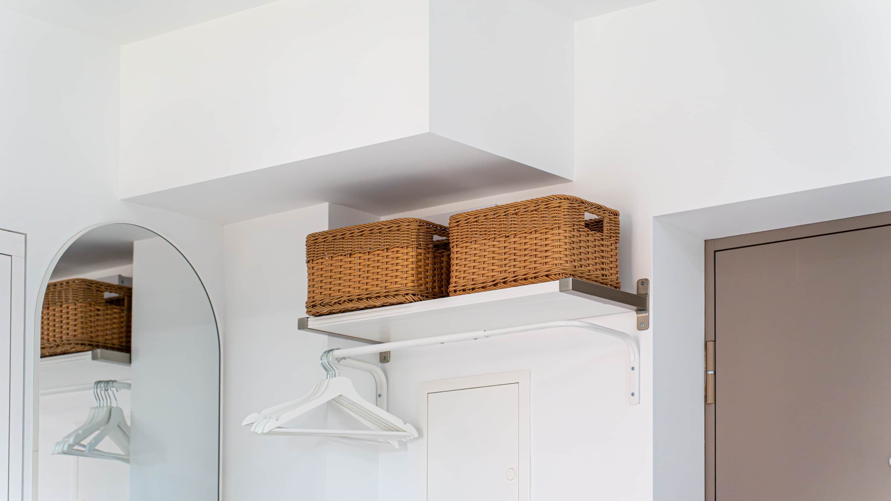 A white interior with a mirrored closet door, a white coat rack, and two wicker baskets on a white shelf.