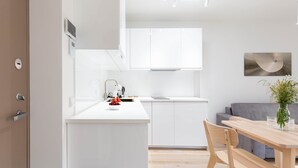 A modern kitchen with white cabinetry, a dining area with a wooden table and chairs, and a grey sofa in the background. - New 2025 Pervalka Neringa Flat White Dunes: AC, Heating, Private Balcony (Pervalka)