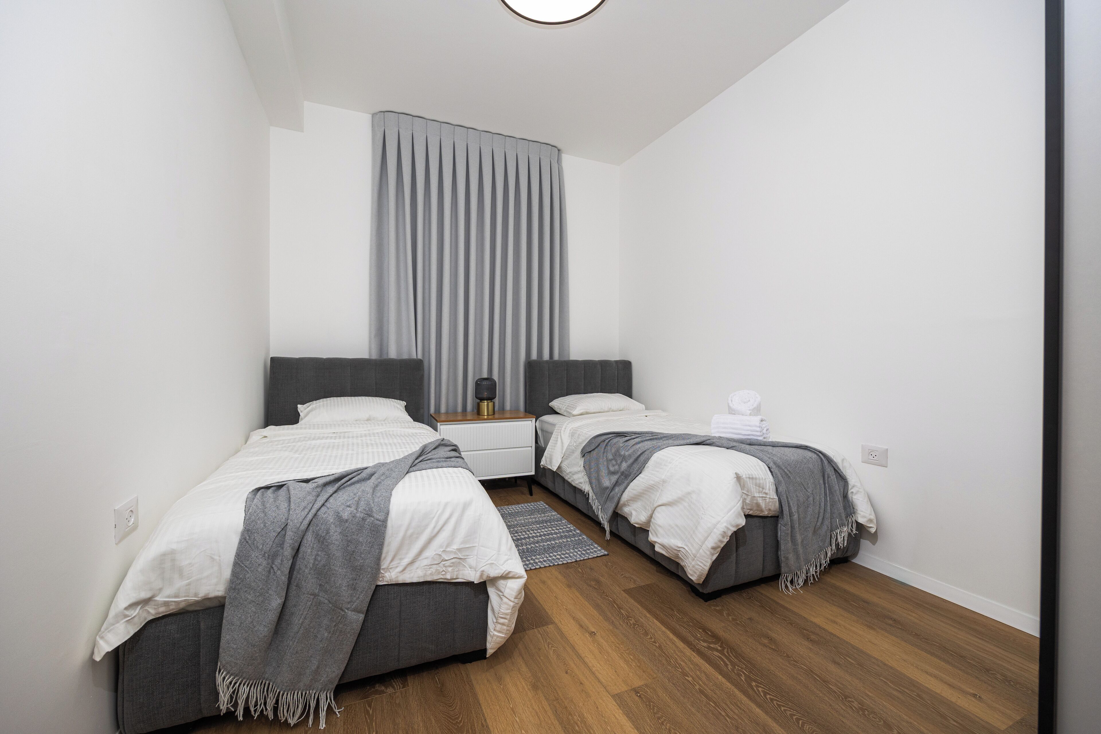 Un dormitorio moderno con dos camas, un ventanal con cortinas, suelo de madera y una mesita de noche con lámpara.