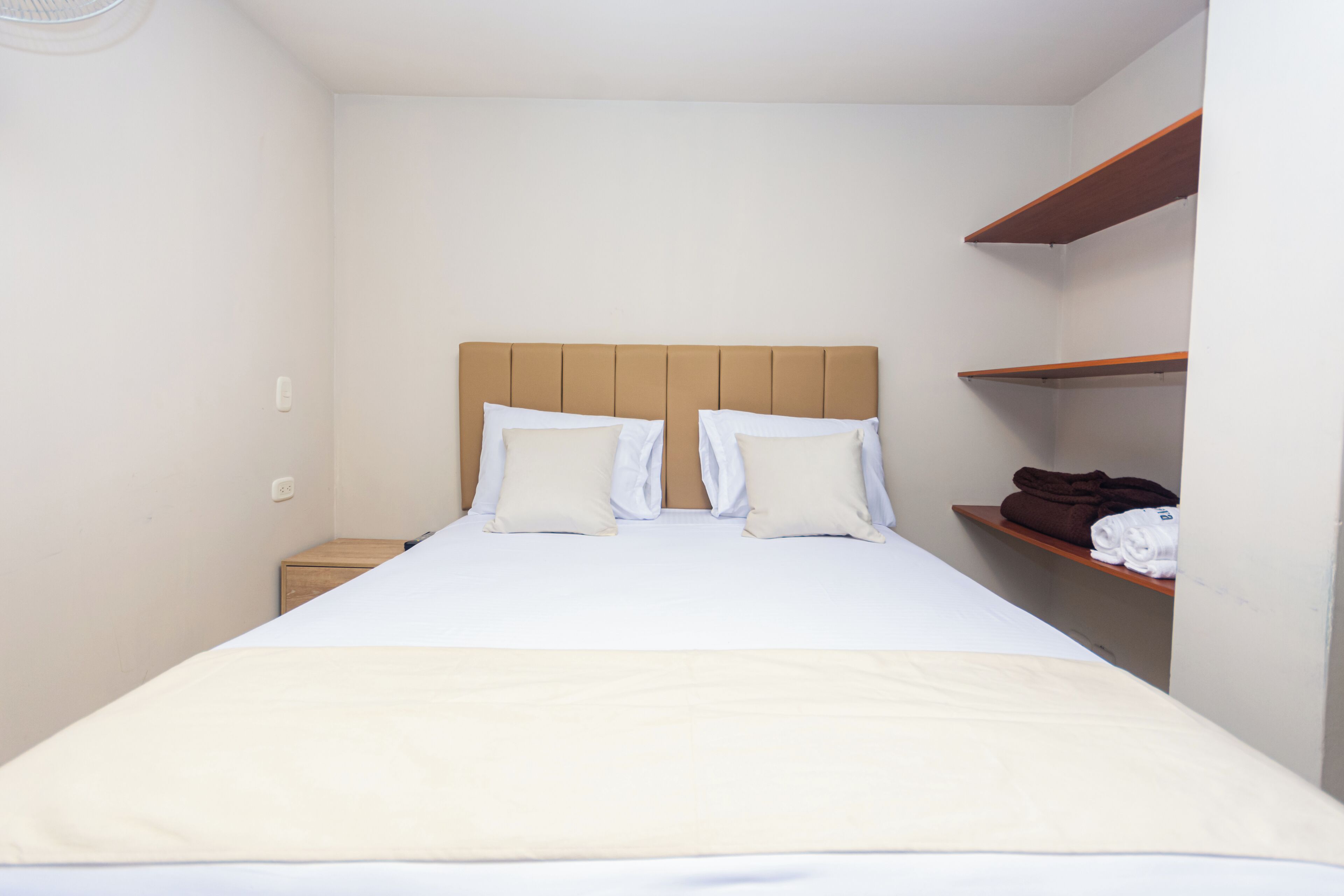 A neatly made bed with a headboard, a wooden nightstand, and a shelf with folded towels.
