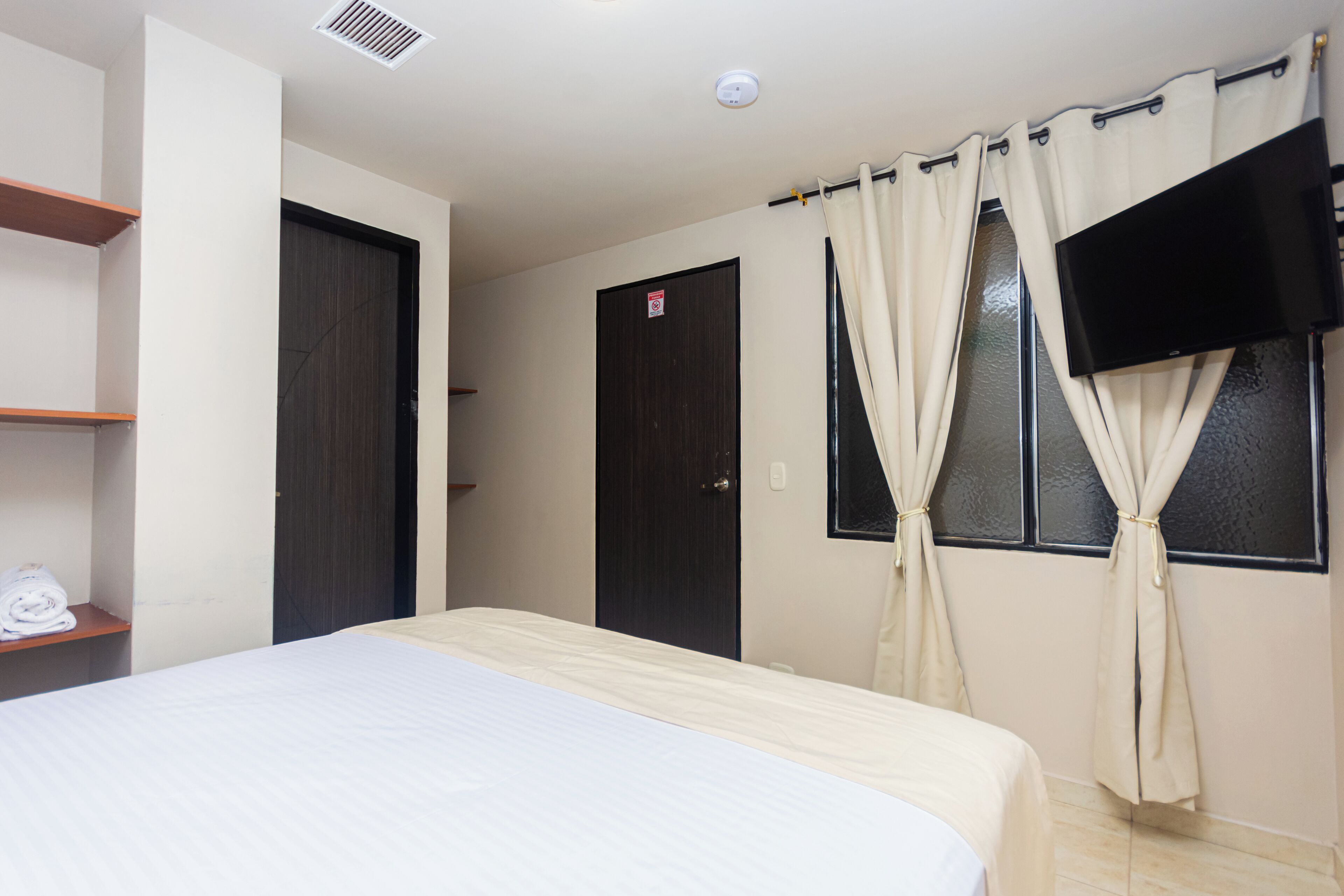 Habitación de hotel con cama, televisor montado en la pared, puerta y ventana con cortinas.