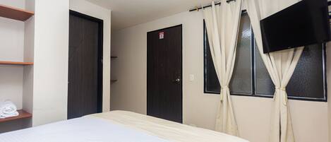 Habitación de hotel con cama, televisor montado en la pared, puerta y ventana con cortinas.