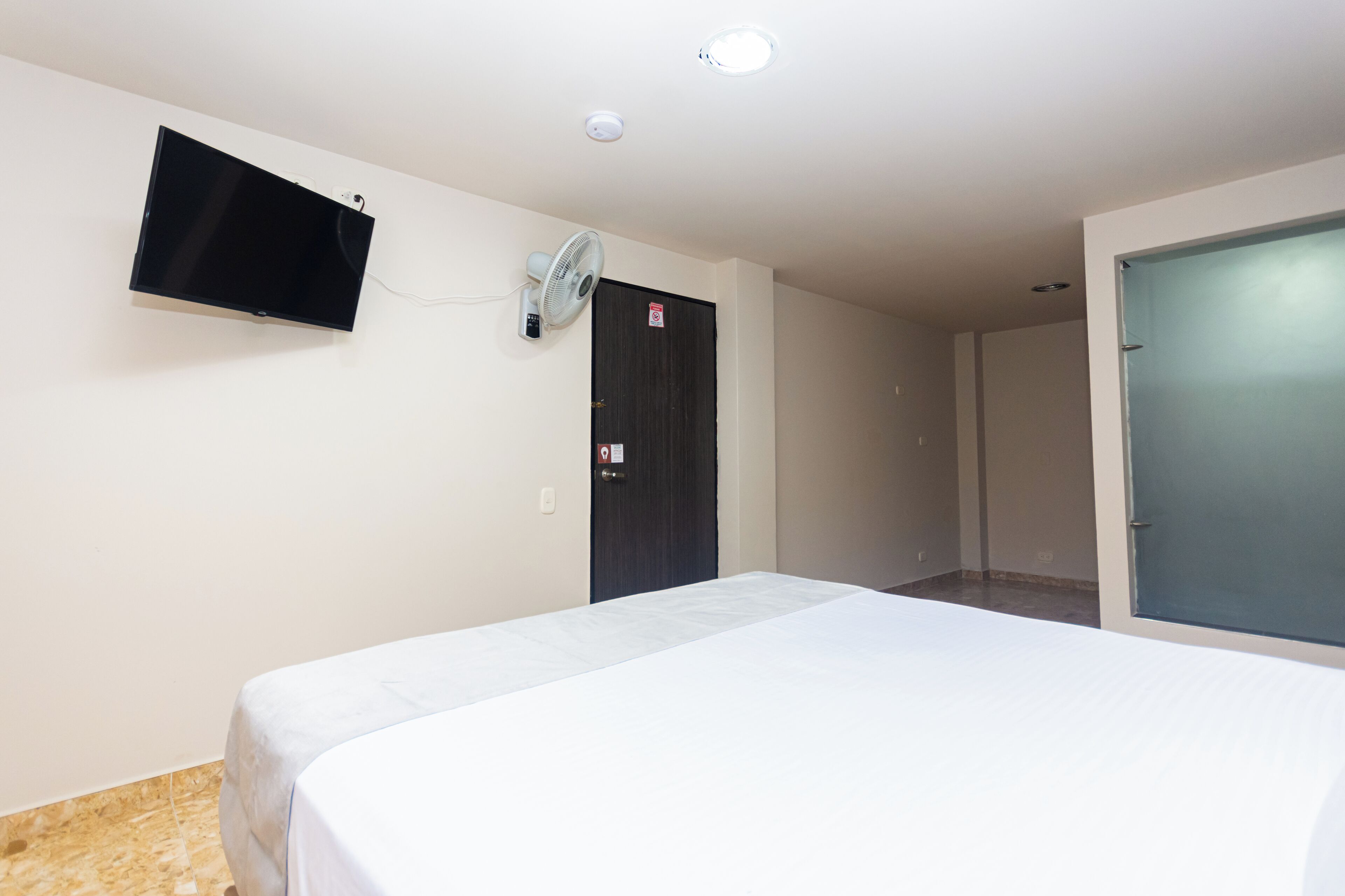 Une chambre d’hôtel avec un lit, un téléviseur fixé au mur, une porte et un ventilateur.