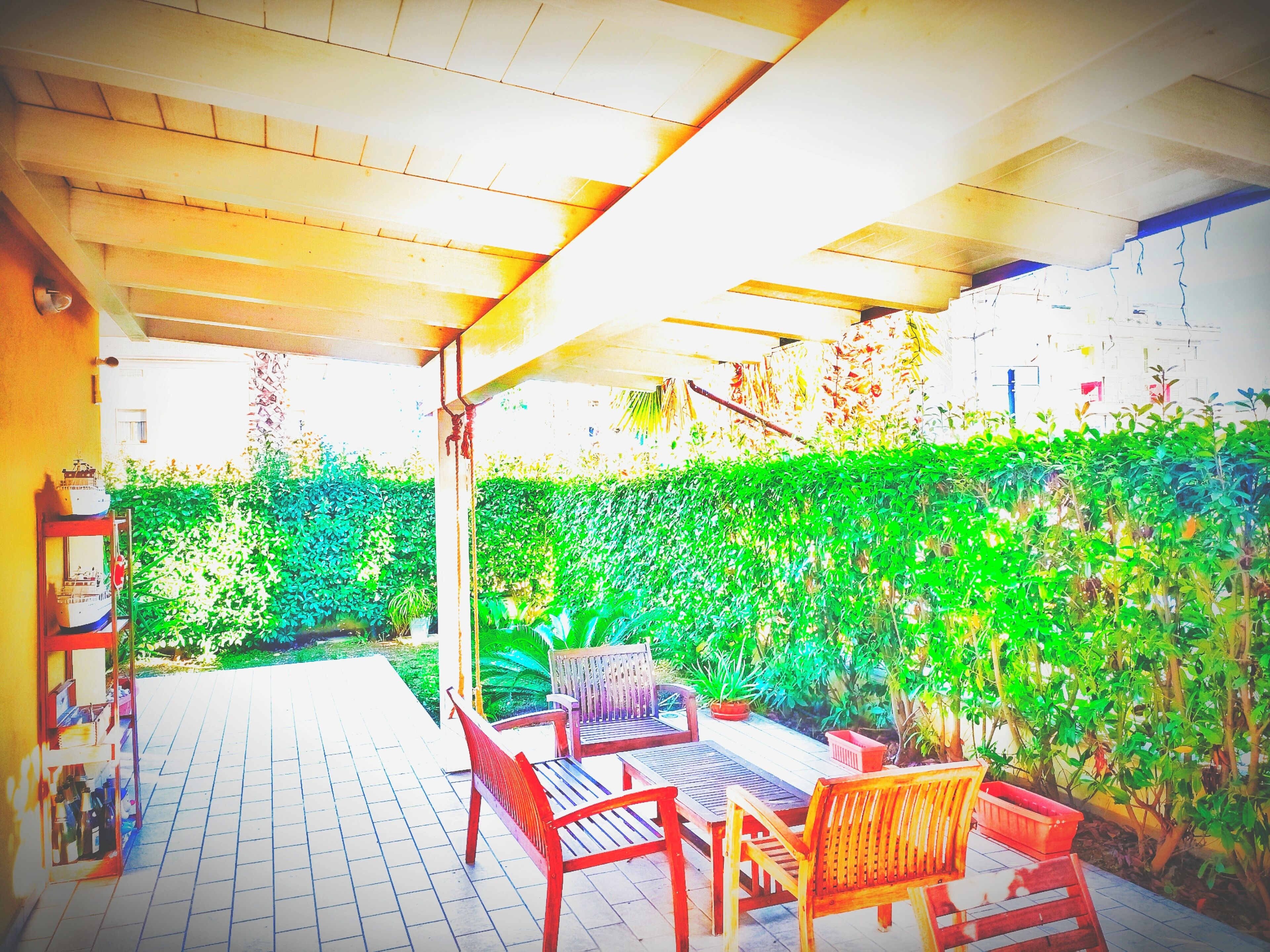 An outdoor patio with a tiled floor, wooden furniture, and a green hedge wall.
