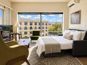 A hotel room with a large bed, a sofa, a TV, a desk, and a view of a building and trees.