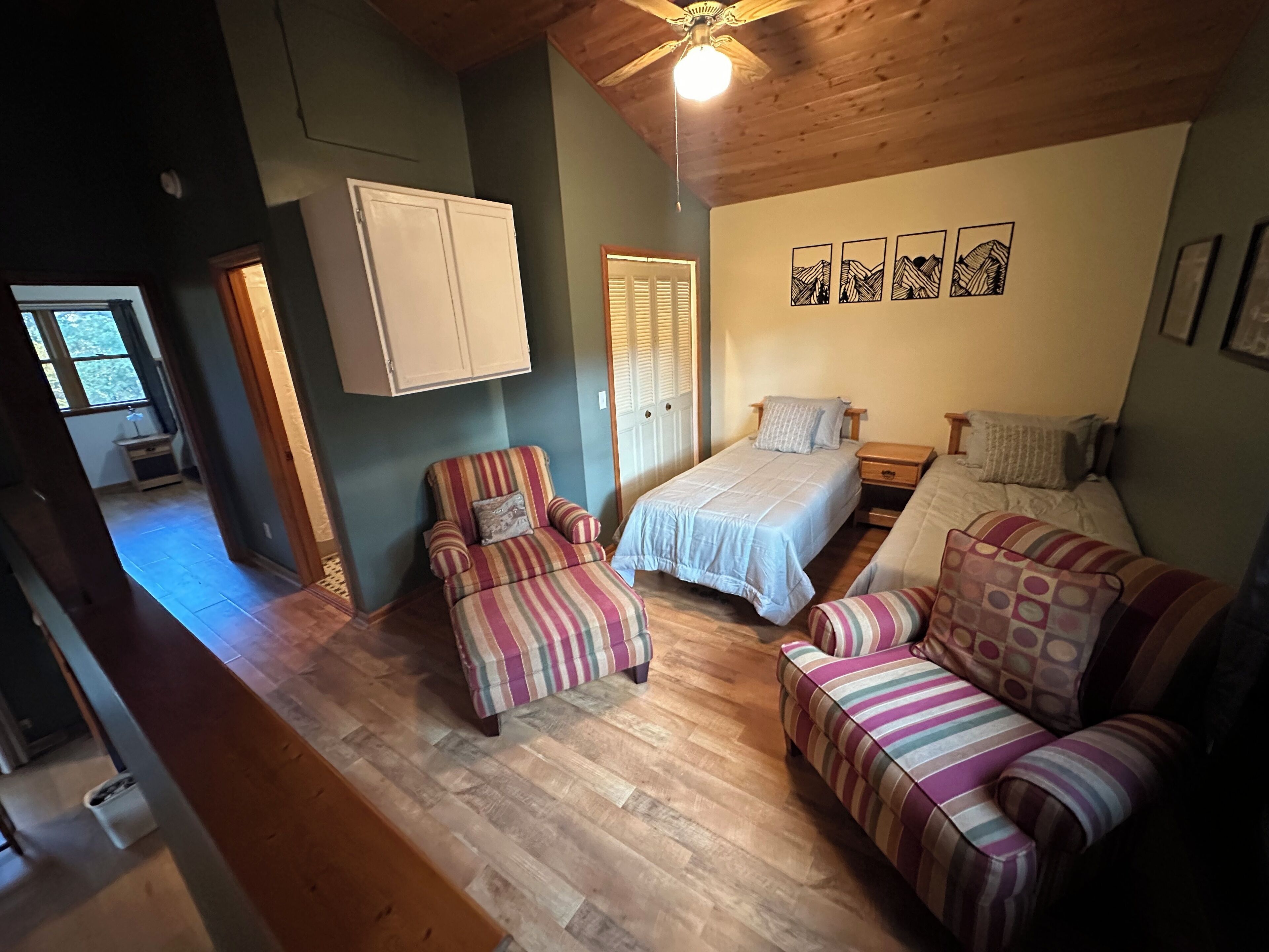 Une pièce confortable avec un lit, deux fauteuils à rayures, un ventilateur de plafond en bois et un mur orné de tableaux encadrés.