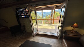 A room with a wooden floor, a sofa, a coffee table, a TV mounted on the wall, and a sliding glass door leading to an outdoor deck with a view of trees and mountains.