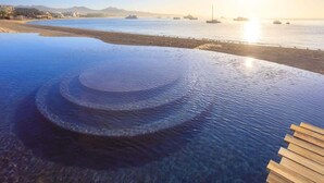 A clear view of an infinity pool with a wooden deck, overlooking a calm sea with boats and a distant shoreline. - Medano Beach Cabo Resort 3 Bedroom  (Cabo San Lucas)