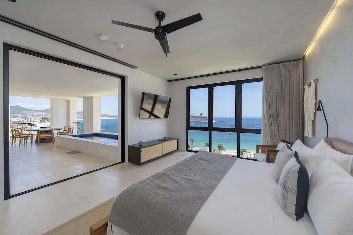 A modern hotel room with a large bed, a ceiling fan, a TV, and a view of the sea.