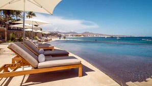 A poolside area with lounge chairs and umbrellas, overlooking a beach with boats and a clear sky. - Medano Beach Cabo Resort 3 Bedroom  (Cabo San Lucas)