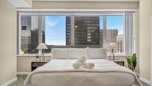 A hotel room with a large bed, two bedside tables, a window with a city view, and a plant on the right side. - LAKE VIEW SkyTower Suite: Downtown ~ King Bed (Cleveland)