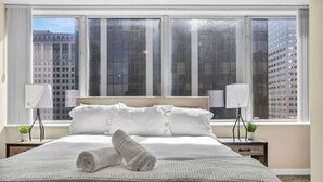 Habitación de hotel con una cama grande, dos mesitas de noche con lámparas y vistas a edificios altos a través de amplias ventanas.