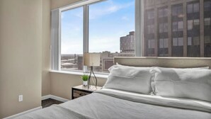 A hotel room with a large window, a bed, a nightstand with a lamp, and a small plant.
