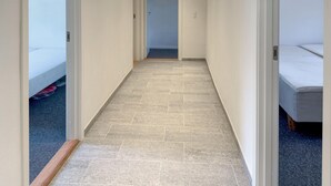 Un couloir étroit, dont le sol est recouvert de carreaux, mène à une pièce où se trouvent un lit et une porte.