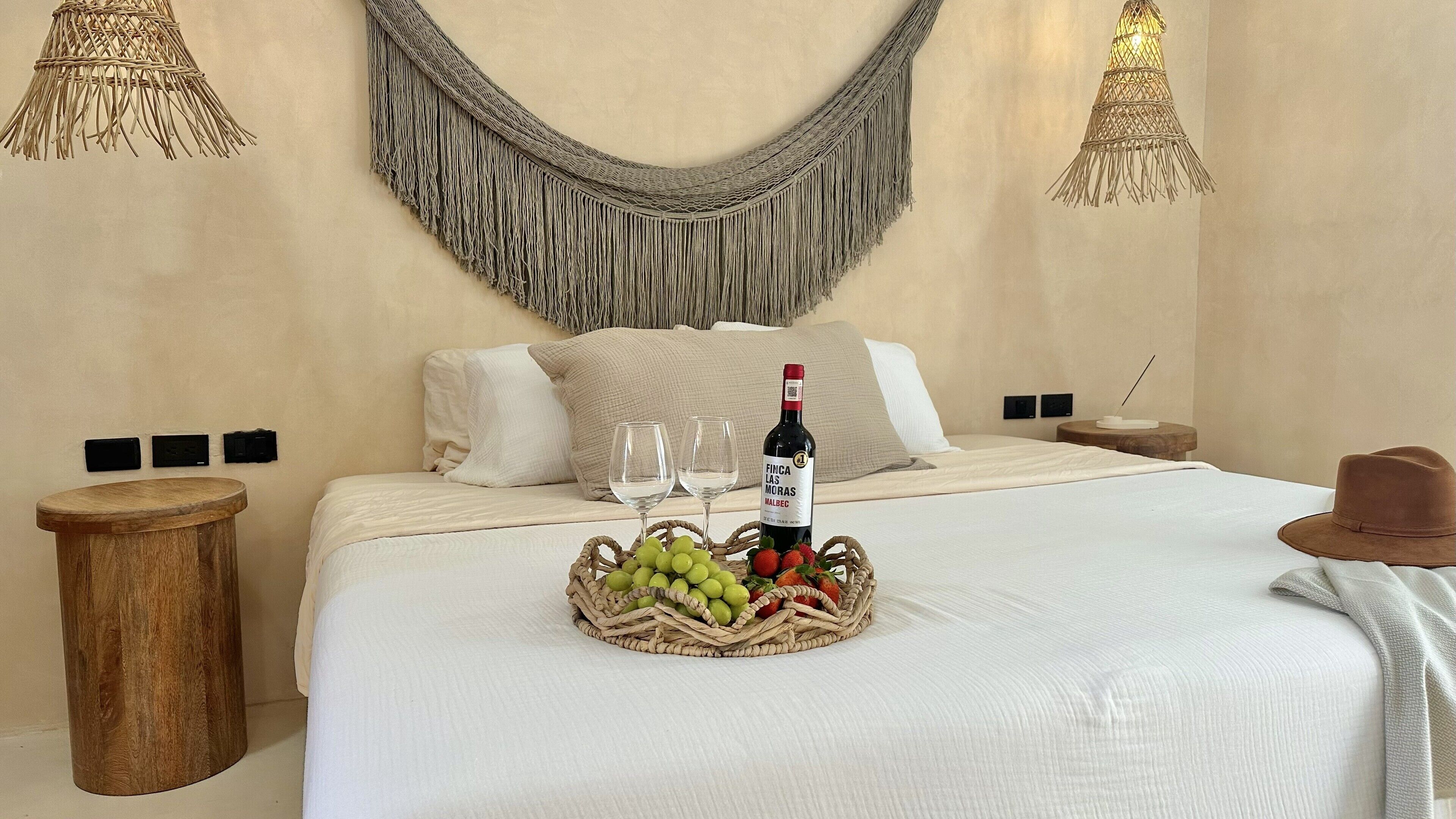 A neatly made bed with a white comforter, two pillows, a bottle of wine, and a basket of fruit on the bed.