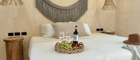 A neatly made bed with a white comforter, two pillows, a bottle of wine, and a basket of fruit on the bed.