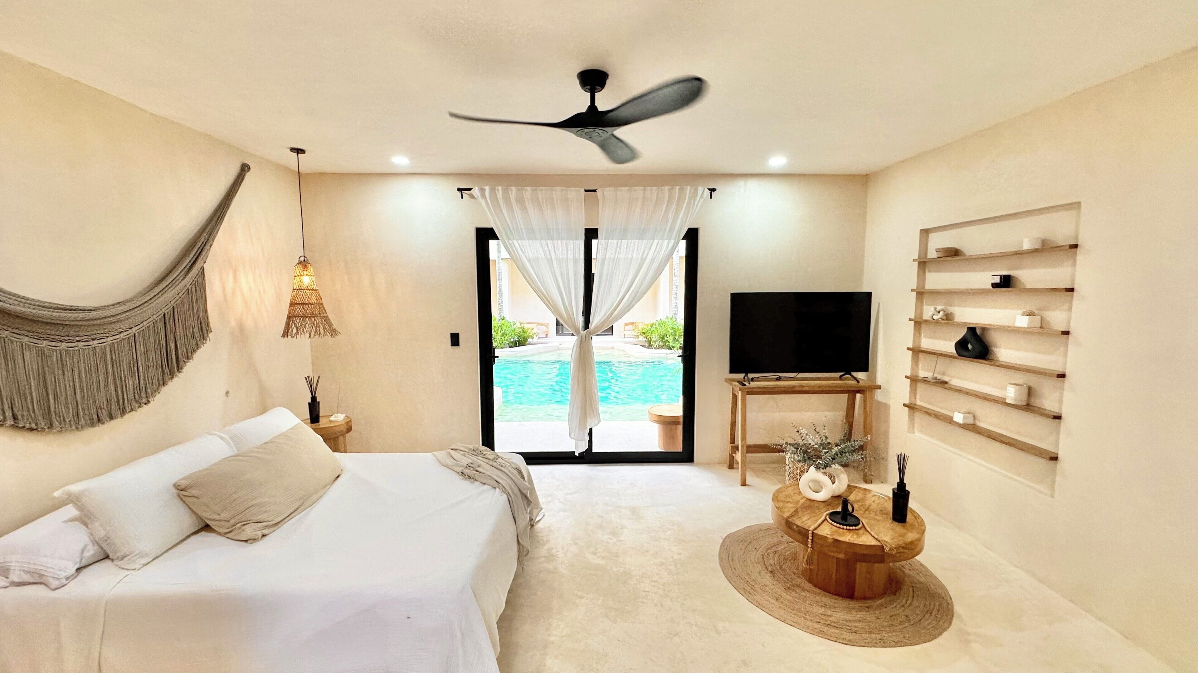 A bedroom with a bed, a TV, a wooden coffee table, a ceiling fan, and a view of a pool.