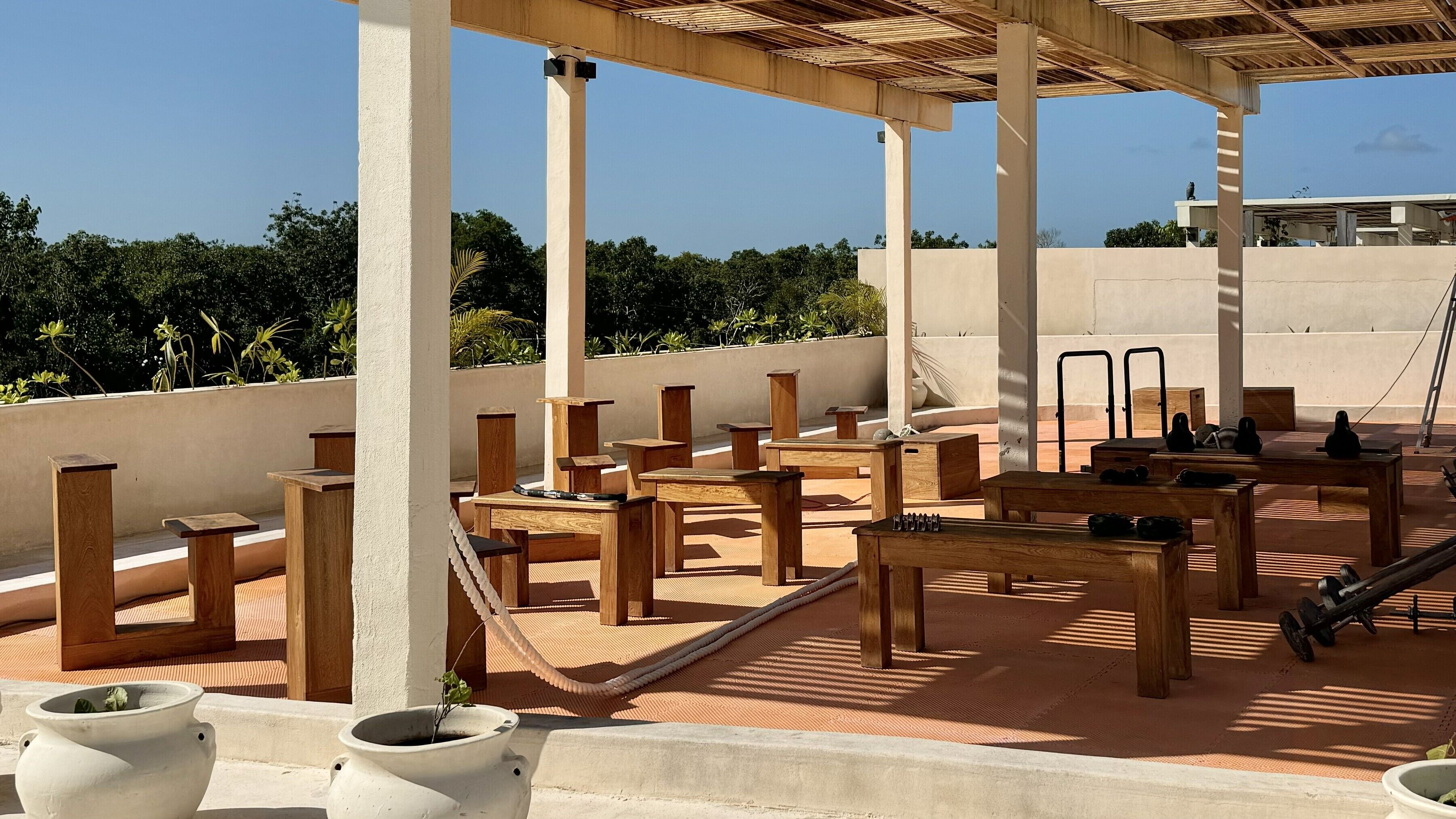 A rooftop terrace with wooden tables and chairs, potted plants, and a view of the surrounding greenery.
