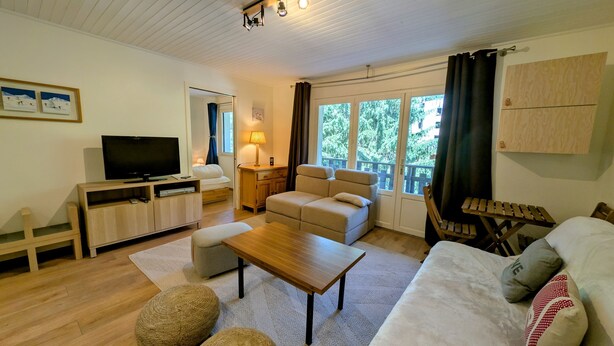 A cozy living room with a sofa, ottoman, wooden coffee table, TV, and a window with curtains.