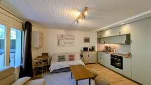 A cozy bedroom with a bed, a small table, and a window. Adjacent is a compact kitchen with a microwave, toaster, and sink.