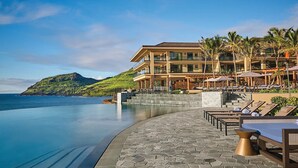 A resort with a pool, lounge chairs, and a view of the ocean and a mountain.