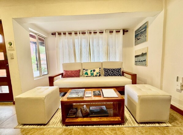 A living room with a sofa set, a coffee table with books, a window with curtains, and a wall with framed pictures. - Island life Duplex great for groups (Puerto Ayora, Santa Cruz Island)