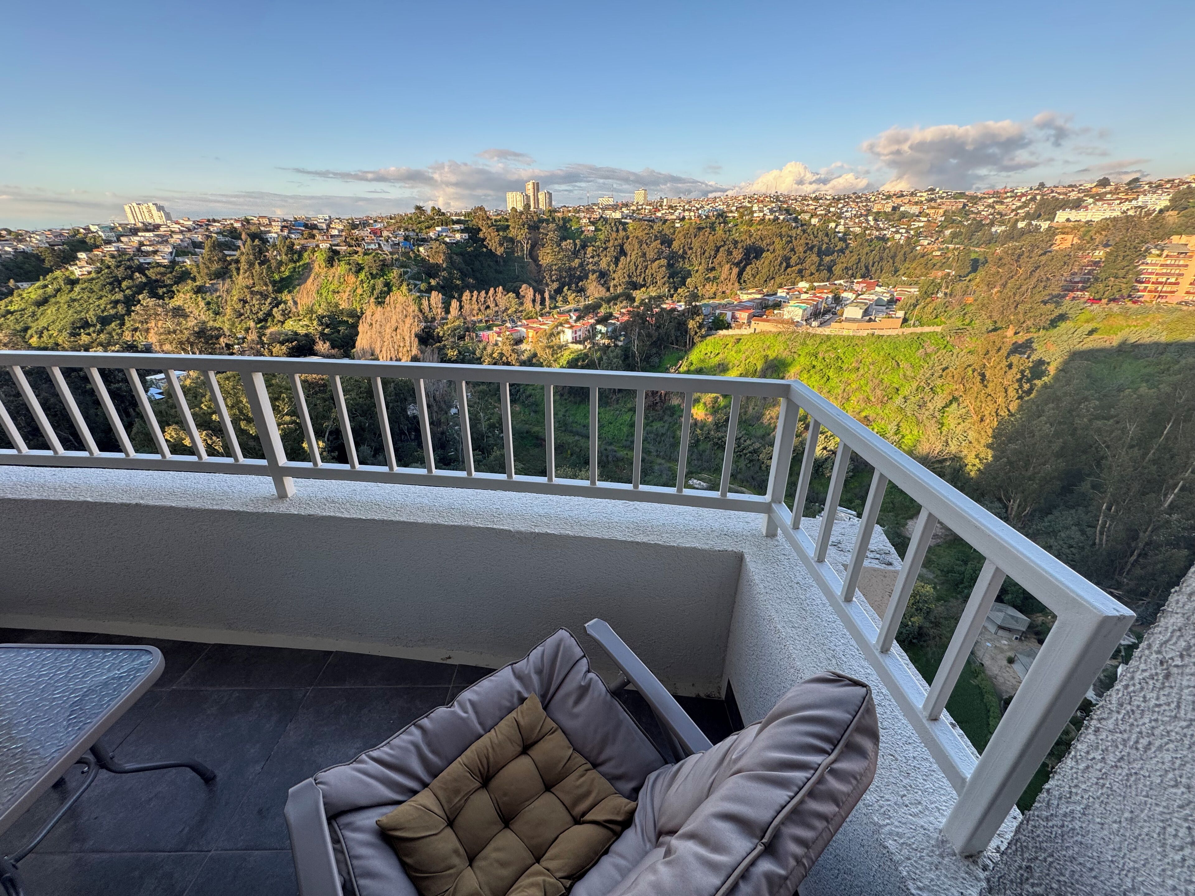 A balcony with a view of a cityscape, a cushioned chair, and a small table.