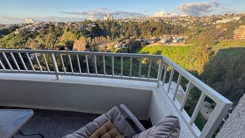A balcony with a view of a cityscape, a cushioned chair, and a small table.
